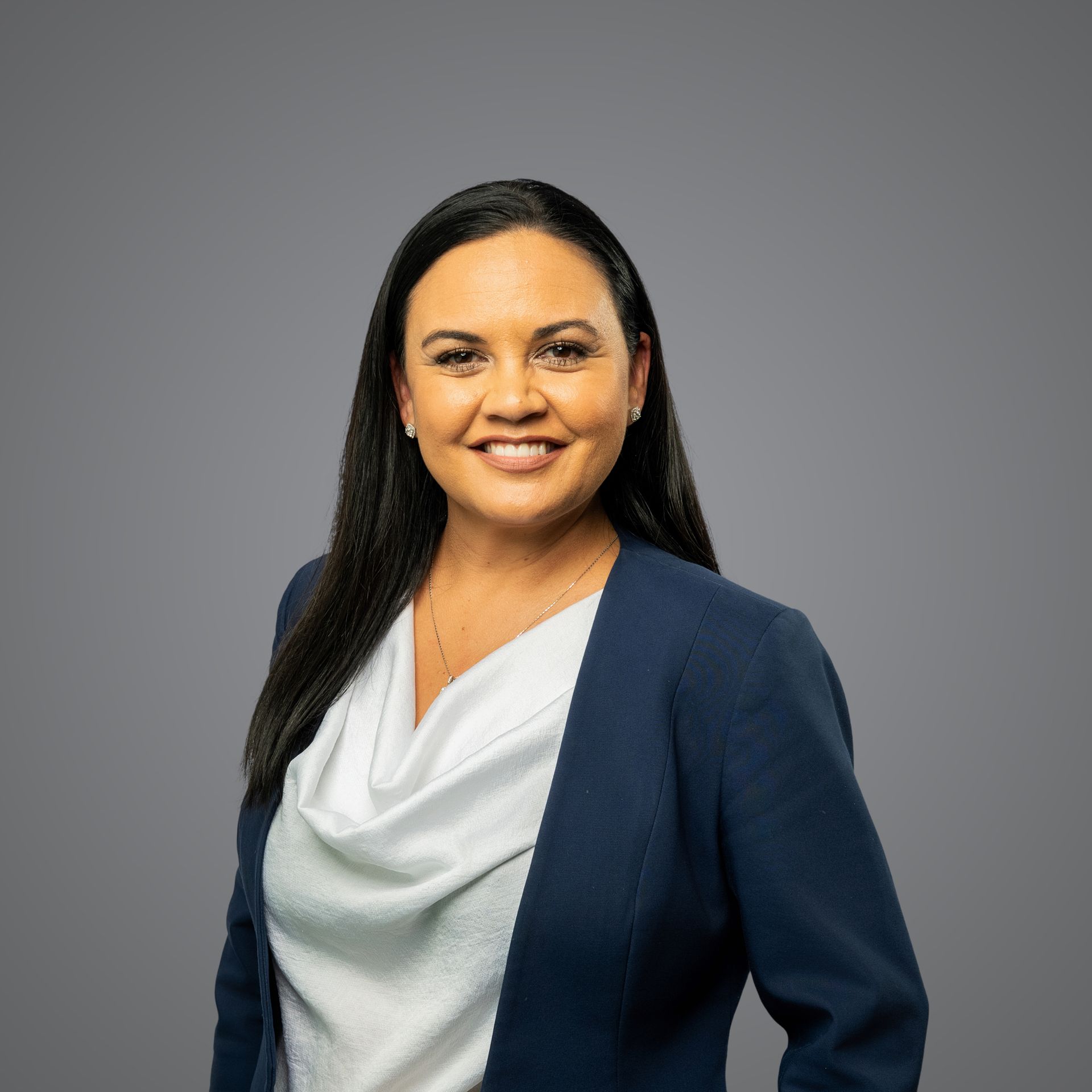 A person wearing a blue blazer and white cowl-neck top smiles against a plain gray background.