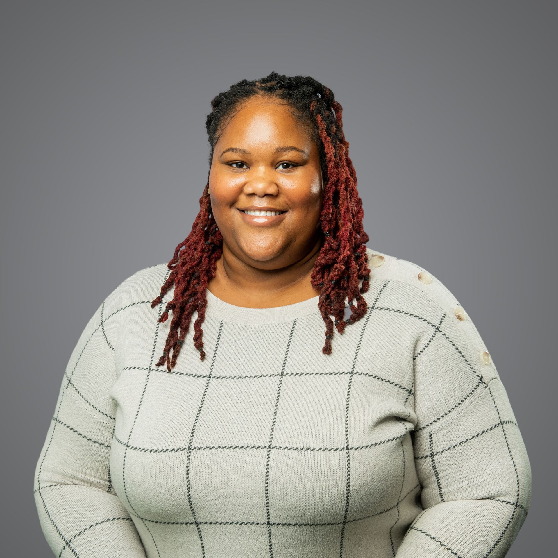 A smiling person with shoulder-length, reddish-brown locs wearing a grey sweater with a black grid pattern.