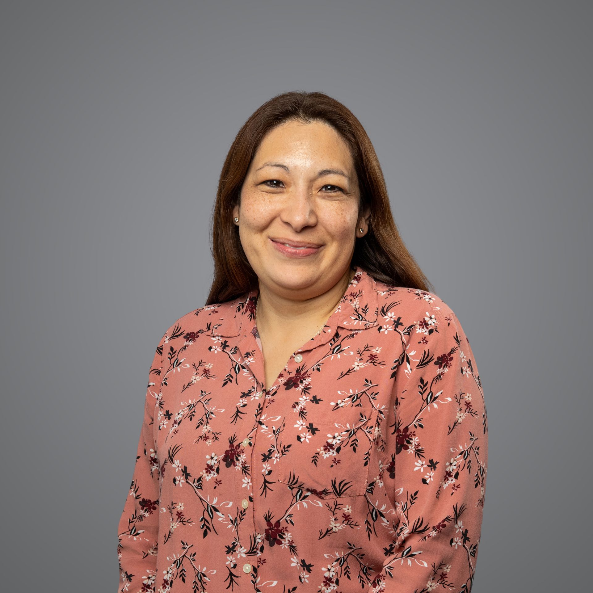 A smiling person with long brown hair wearing a pink floral blouse, centered against a plain gray background.