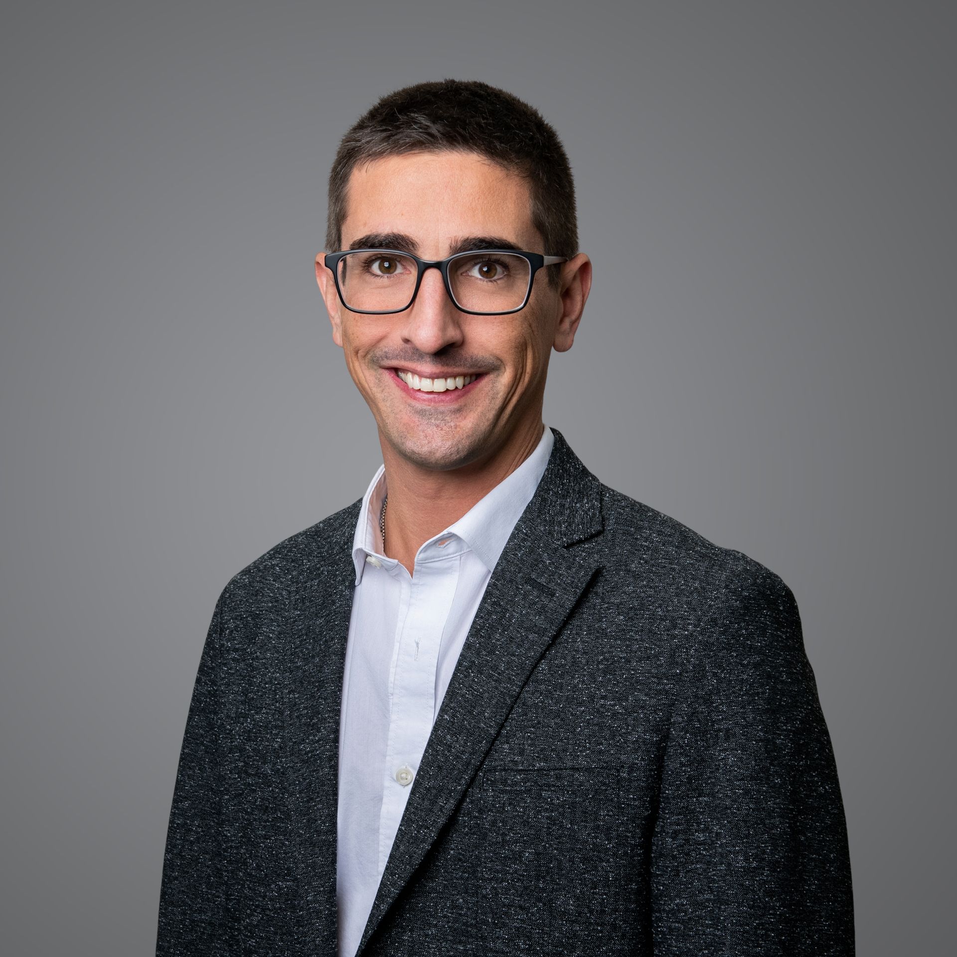 A smiling professional with glasses, wearing a white button-down shirt and a textured charcoal blazer against a gray wall.