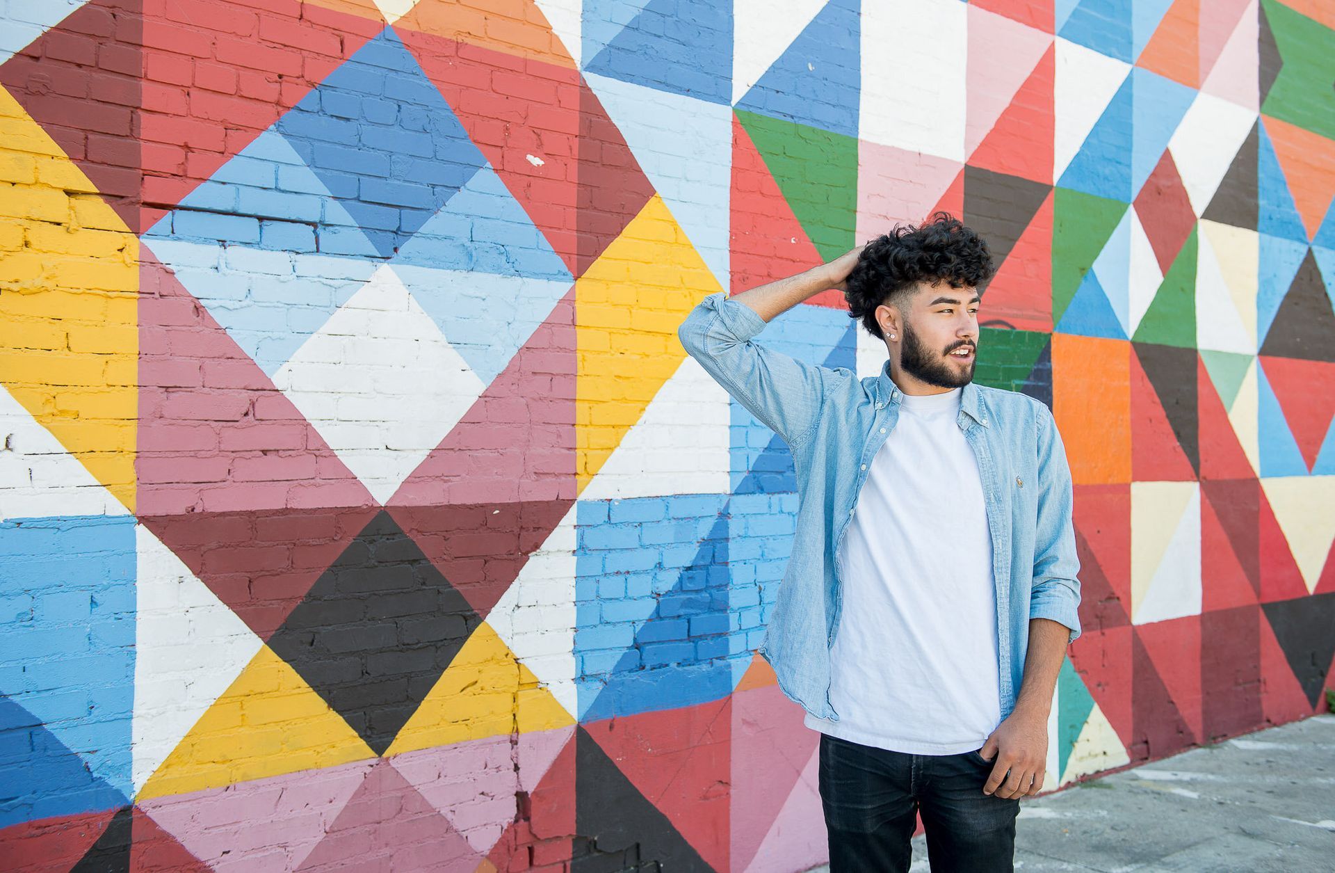 A person stands in front of a colorful, geometric mural, one hand resting on their head, looking away from the camera.