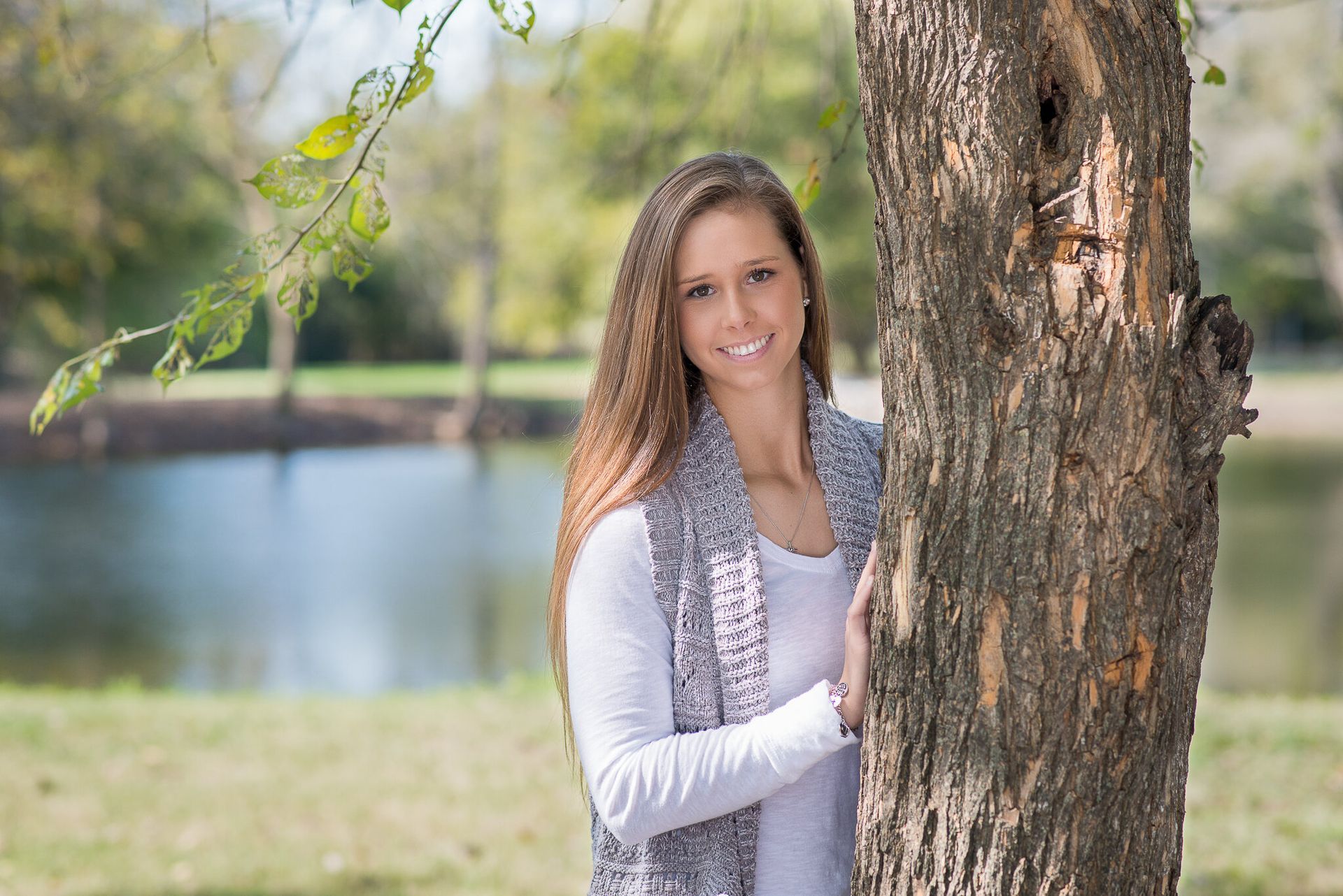A person with long hair smiles while leaning against a tree in a park with a lake in the background.