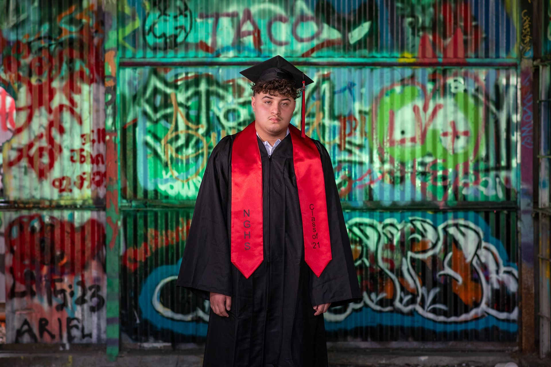 A student in a graduation cap and gown with a red stole standing in front of a wall covered in colorful graffiti.