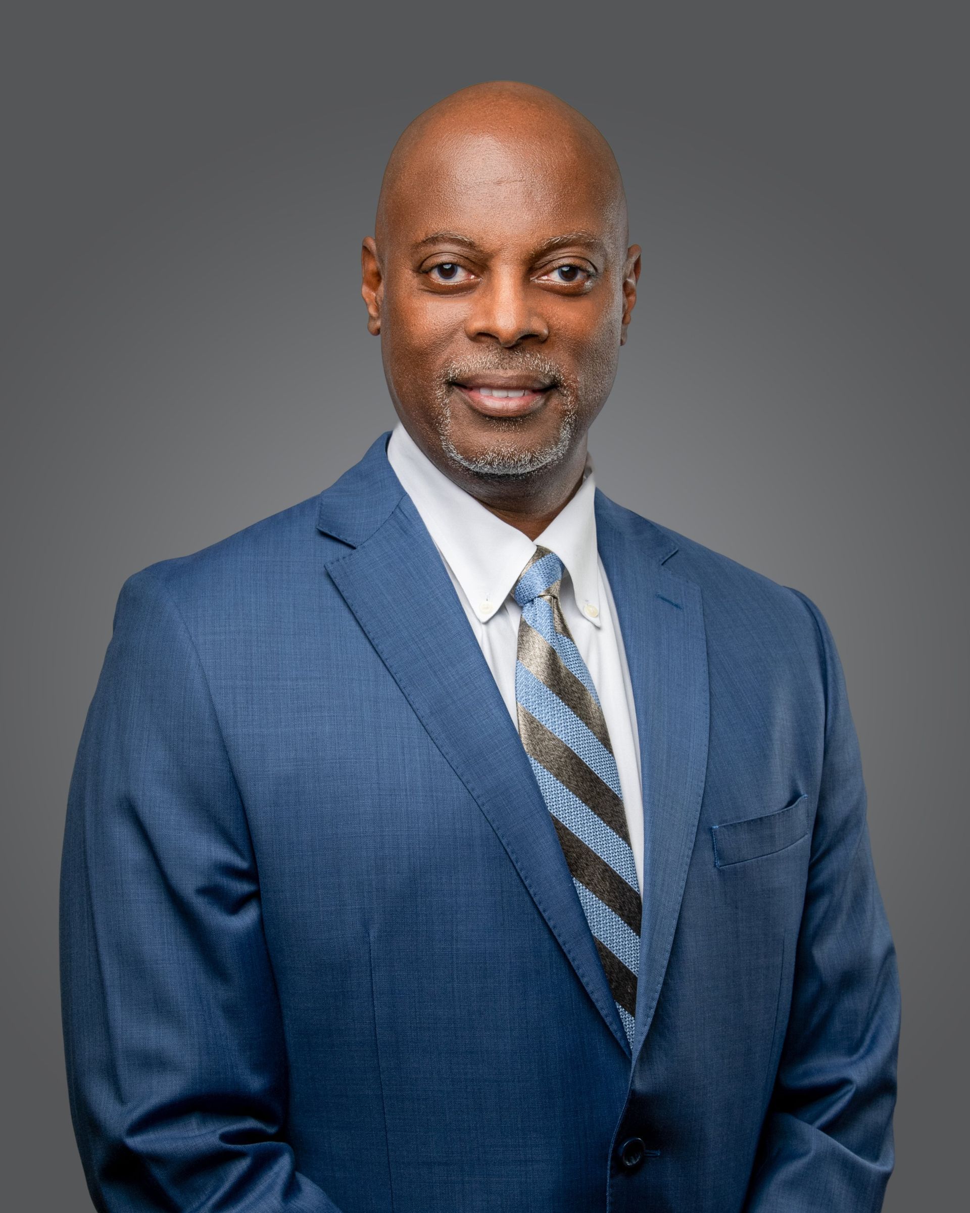 A professional headshot of a person wearing a blue suit, white shirt, and a blue and grey patterned tie against a grey.