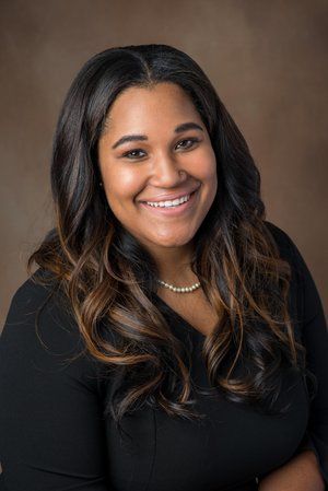 A person with long, dark, wavy hair smiling, wearing a black top and a pearl necklace against a solid brown background.