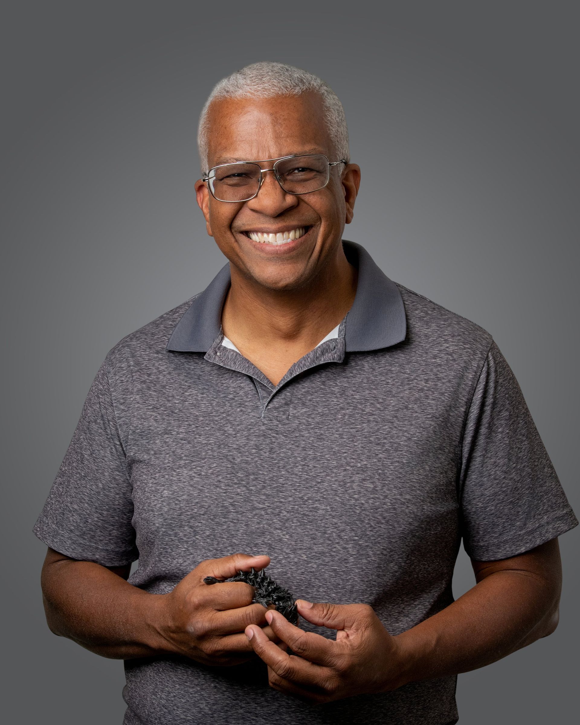 A smiling person wearing glasses and a gray polo shirt, holding a small black object in their hands against a gray background.