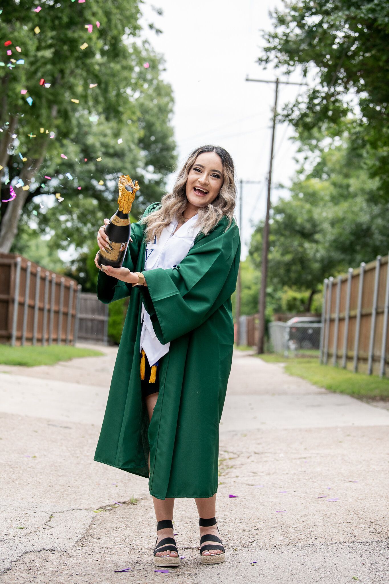 A graduate in a green robe stands on a path, joyfully spraying a bottle of champagne while confetti falls around them.