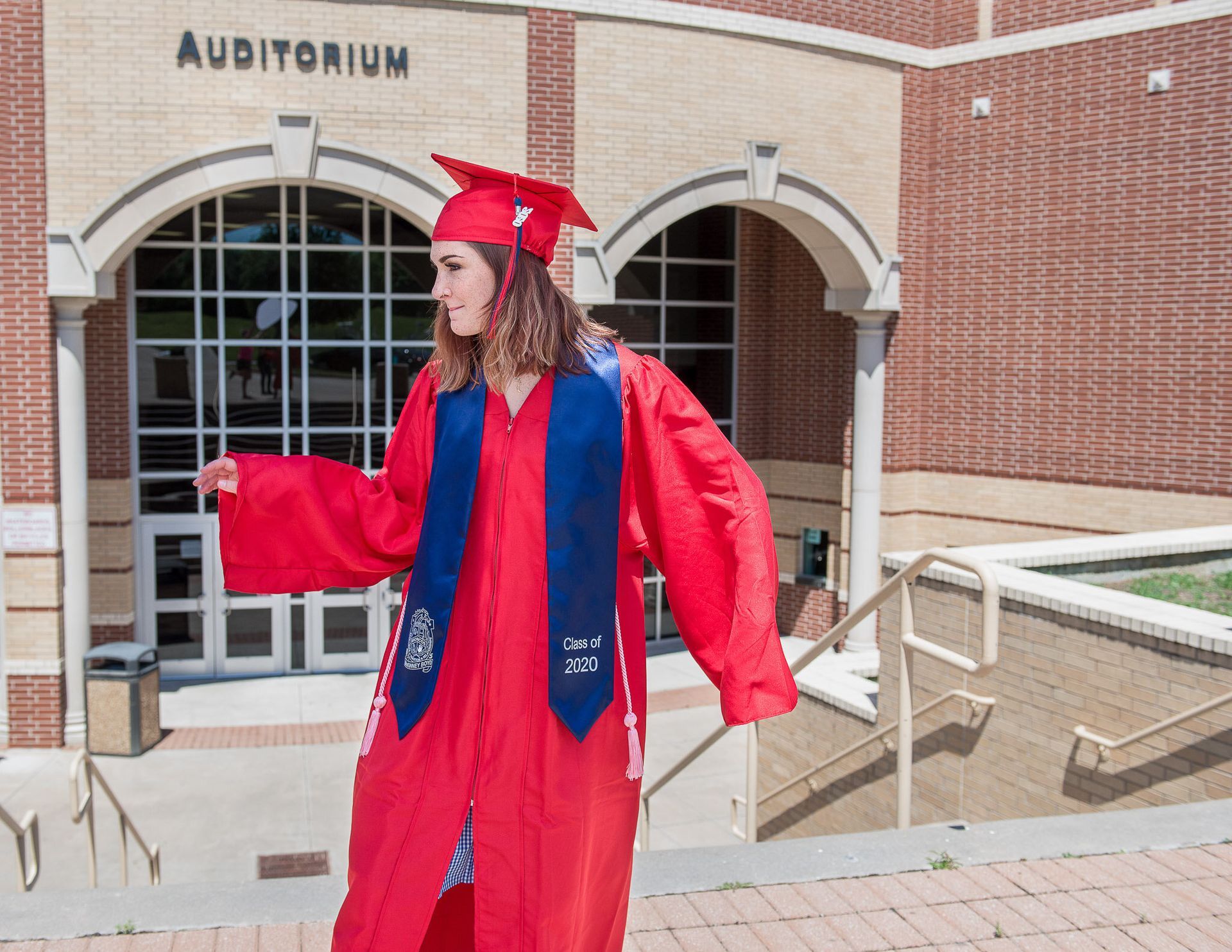 A graduate in a red cap and gown with a blue stole stands outside an auditorium entrance, looking to the side.