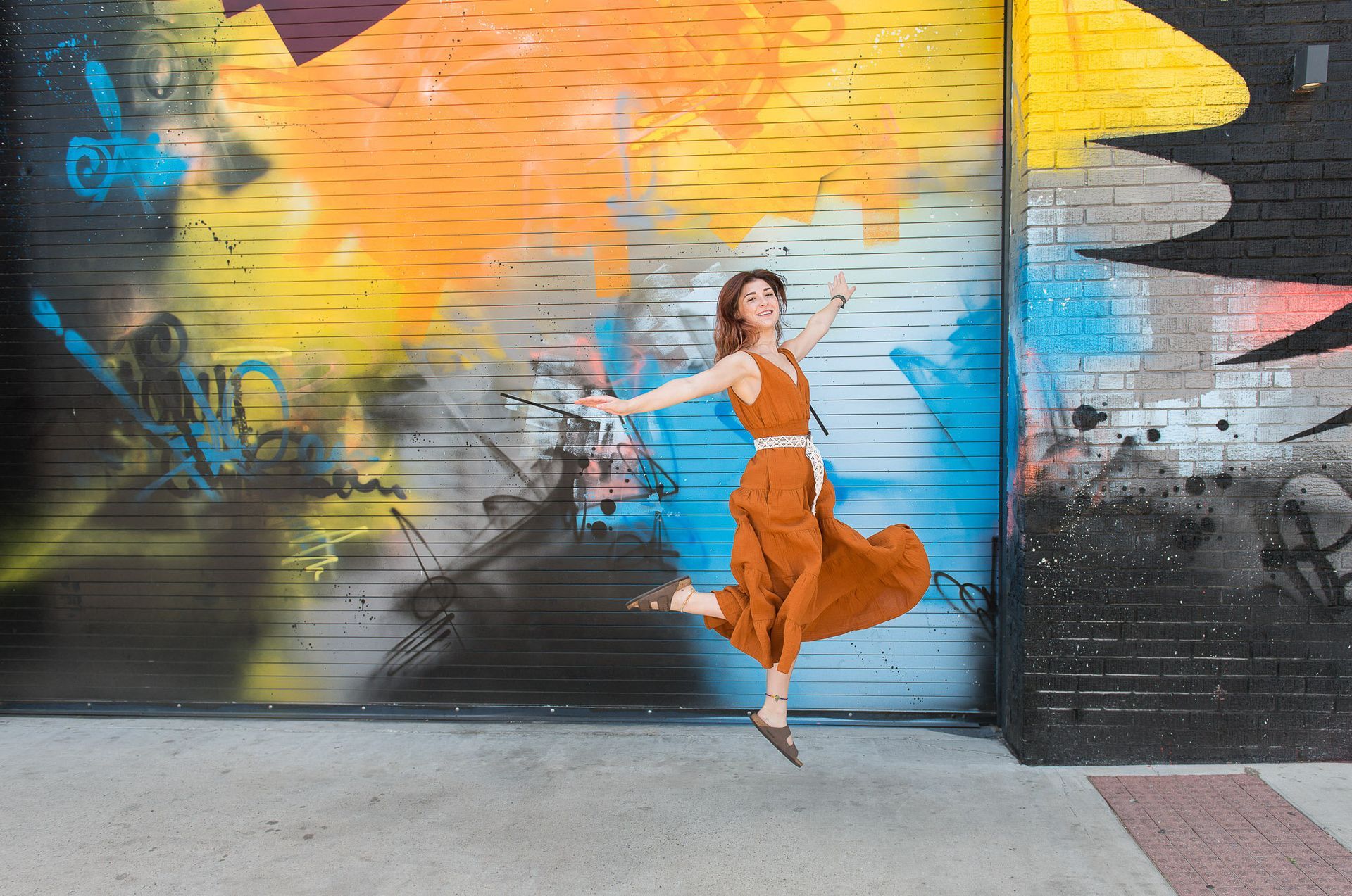 A person in a rust-colored dress jumps in front of a vibrant, multicolored graffiti wall.