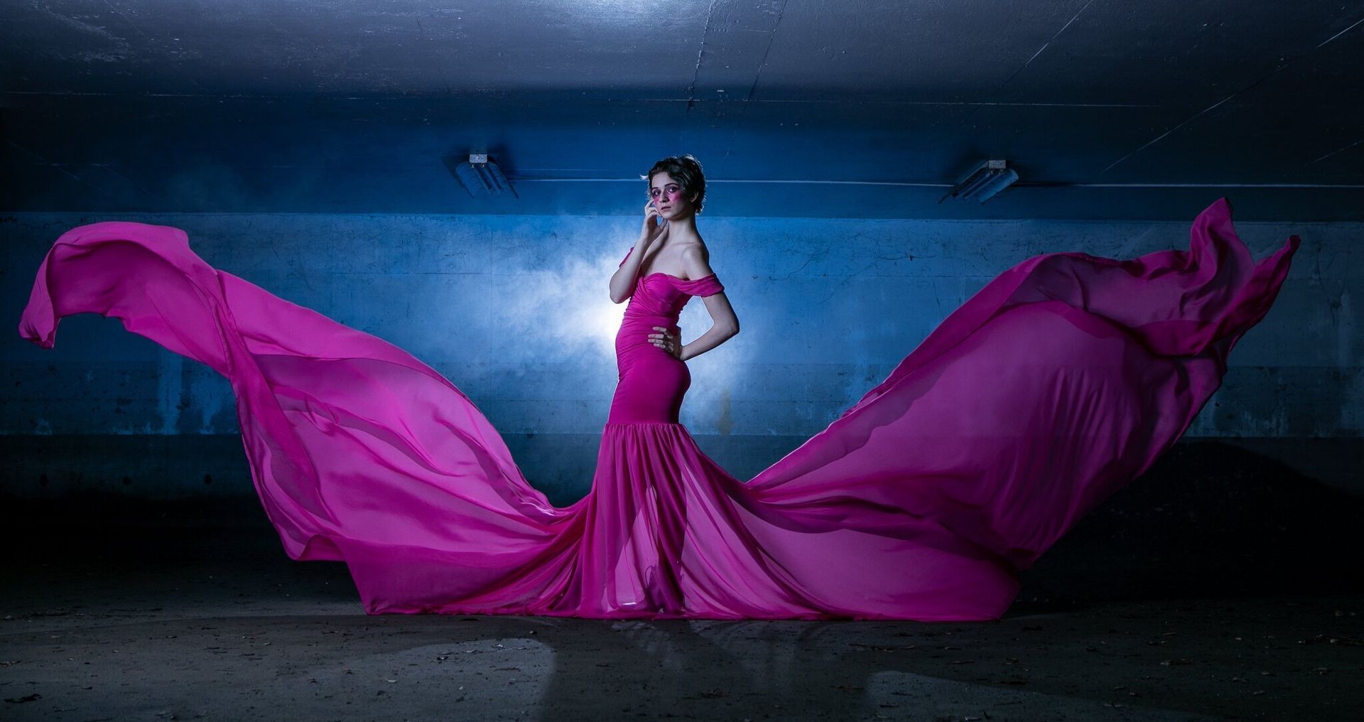 A person in a bright pink flowing gown stands in a dark, dimly lit space as the dress fabric billows out on both sides.