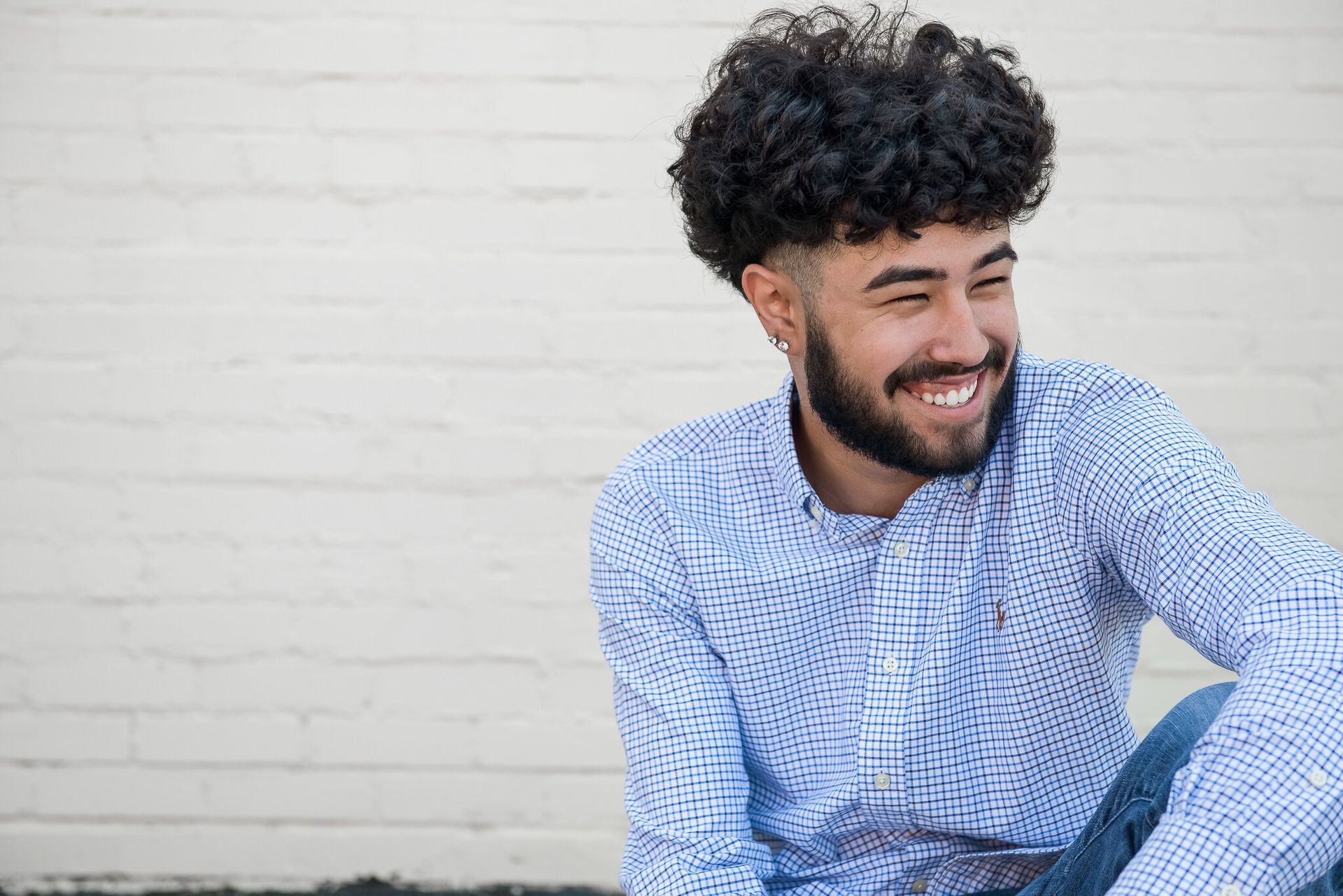 A smiling person with curly hair and a beard, wearing a patterned blue button-down shirt against a white brick wall.