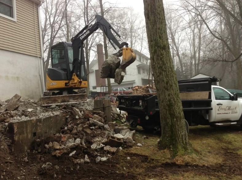 A John Deere excavator demolishes a concrete structure next to a white pickup truck, clearing rubble.
