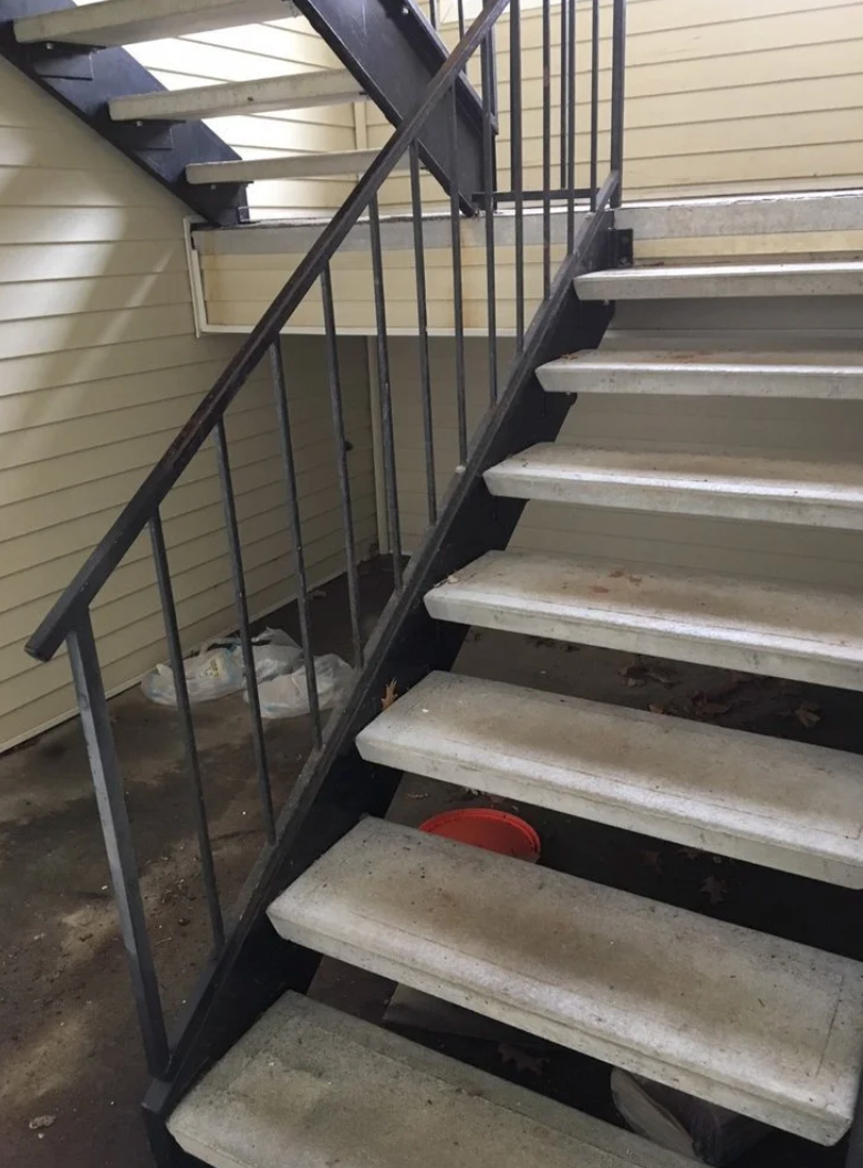 Outdoor stairwell with metal railing. Concrete steps lead upward. Beige siding, dark metal, and a red object visible.