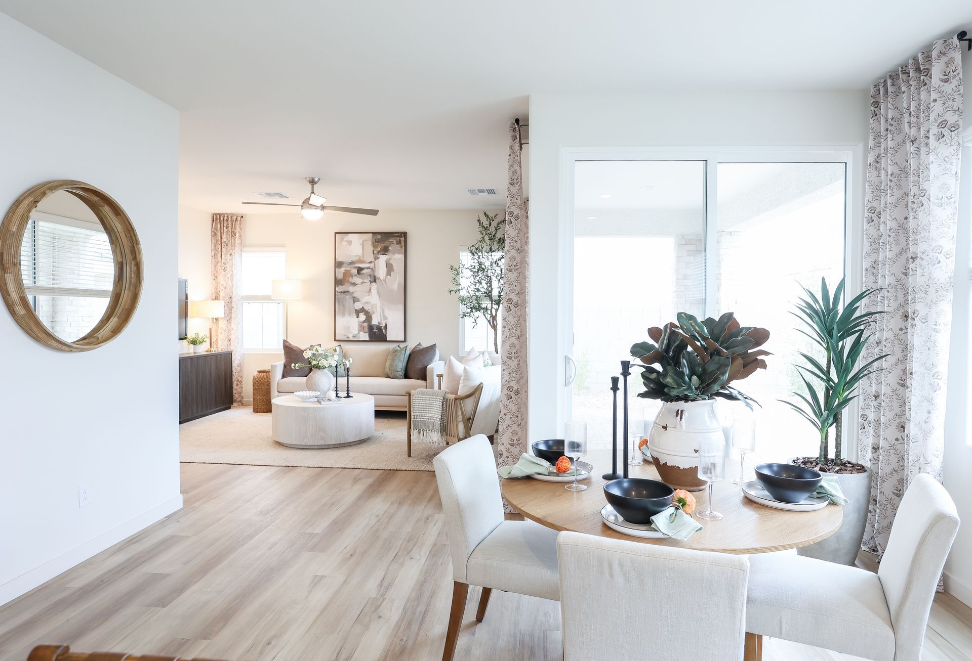 Light-filled dining area with round table, chairs, and view into living room with sofa, artwork, and large window.