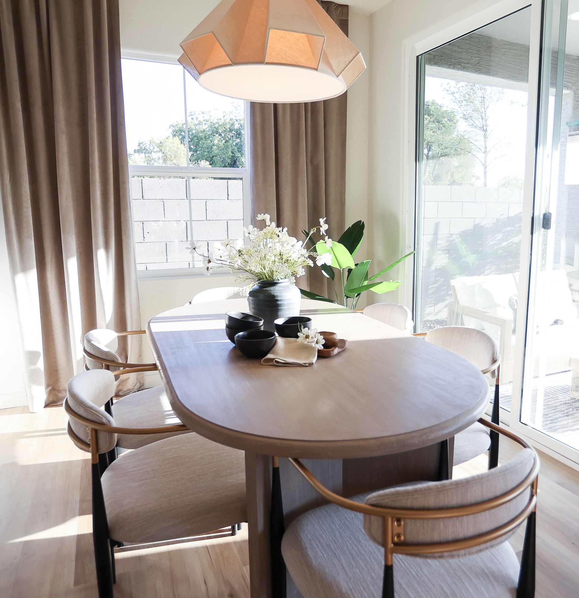 Dining area with a light wood table, beige chairs, and floral centerpiece, with large windows and a geometric pendant light.
