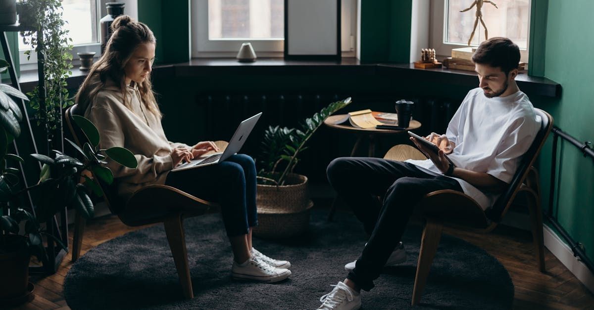 A man and a woman are sitting in chairs in a living room using laptops.