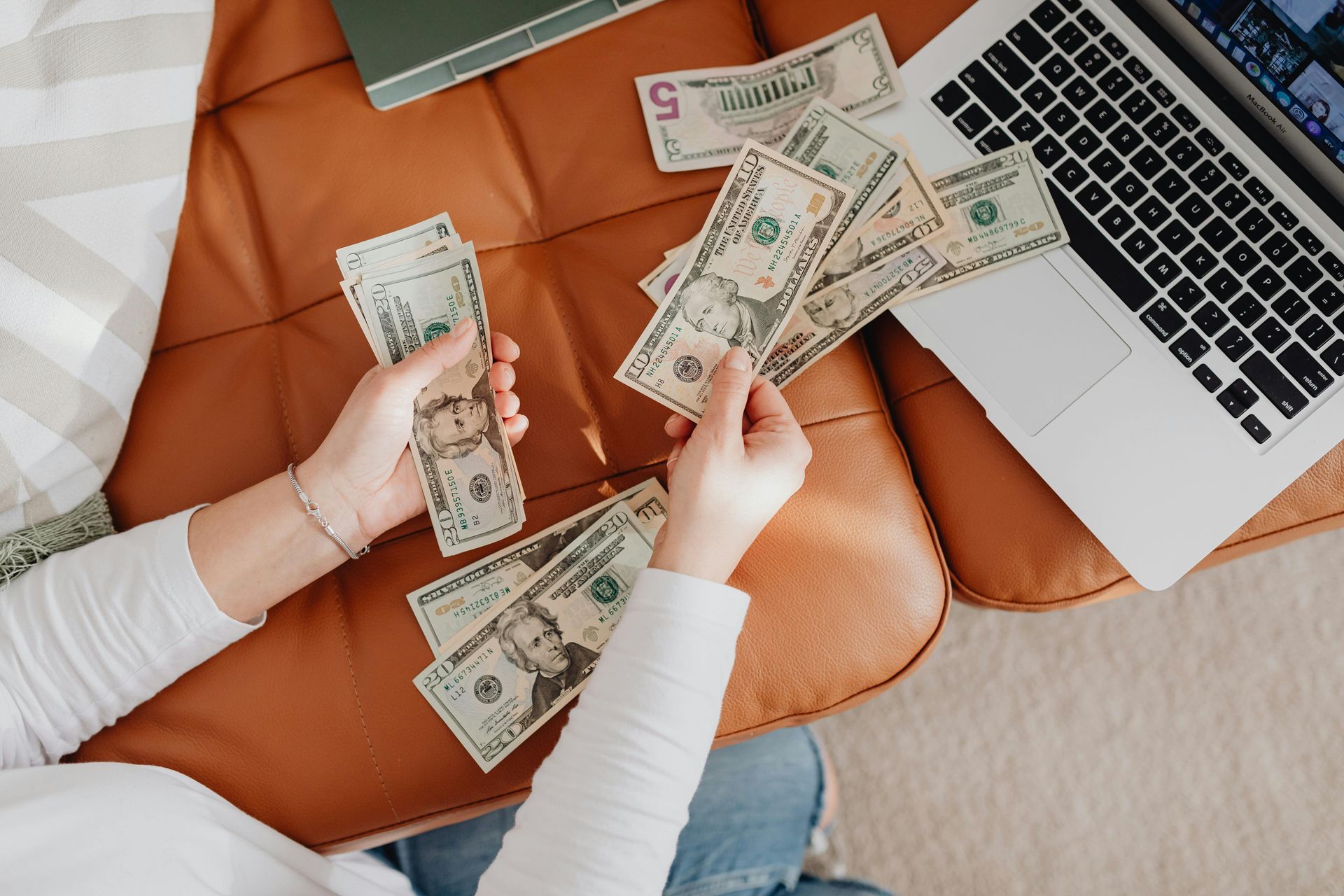 A woman is sitting on a couch counting money next to a laptop.