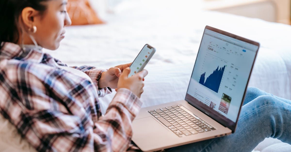 A woman is sitting on a bed using a laptop and a cell phone.