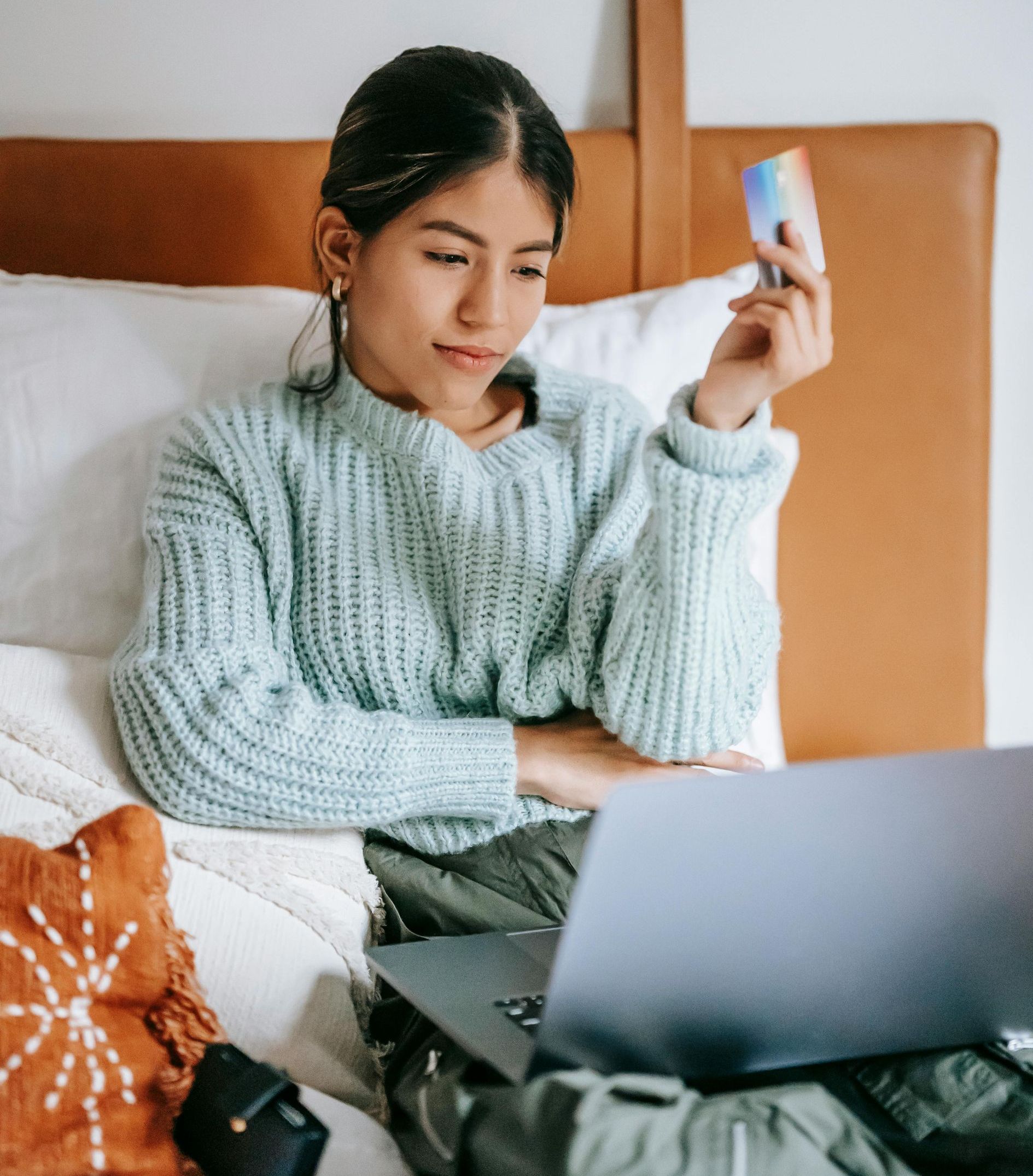 A woman is sitting on a bed using a laptop and holding a credit card.