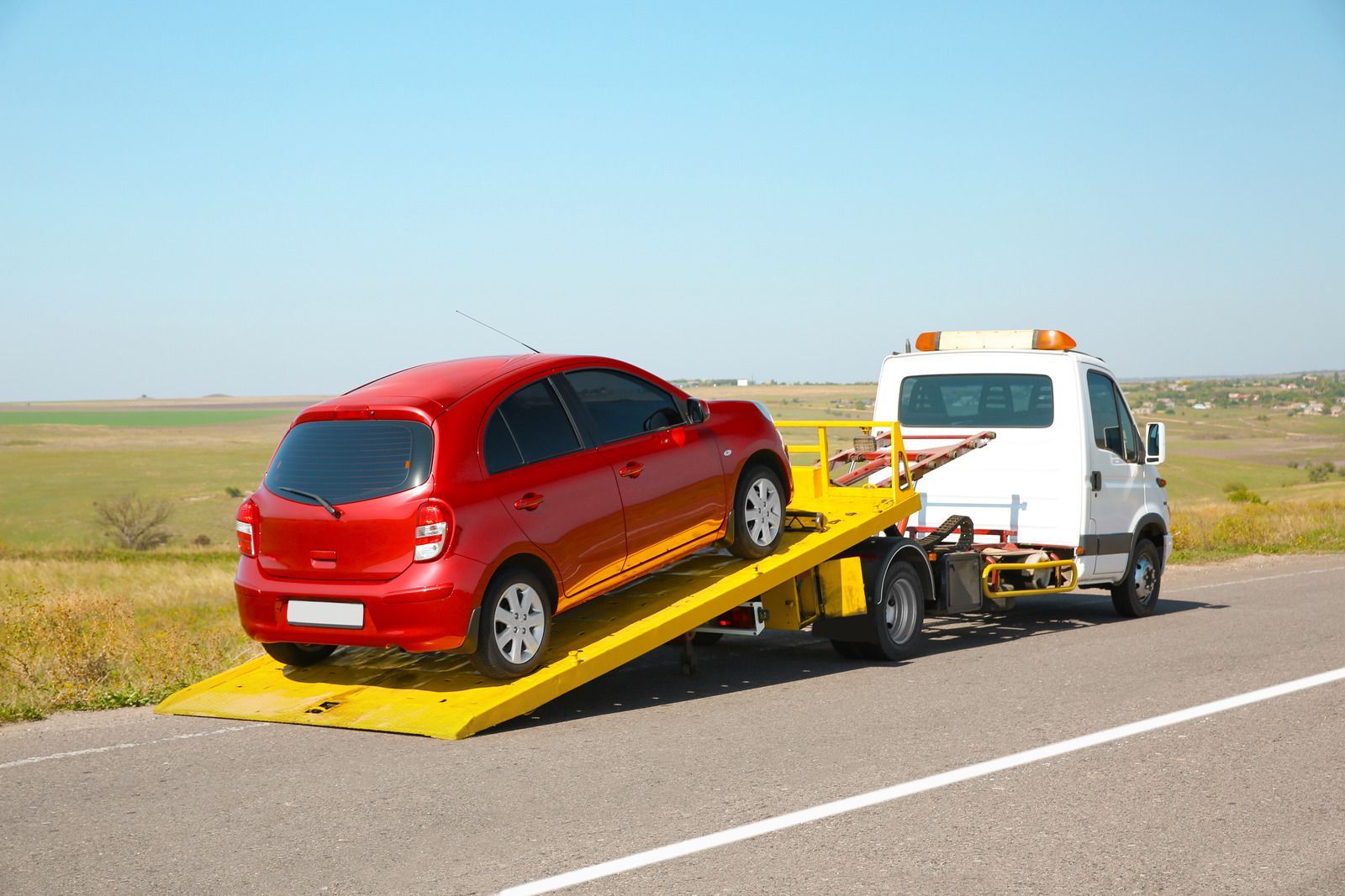 A red car is being towed by a tow truck on the side of the road.