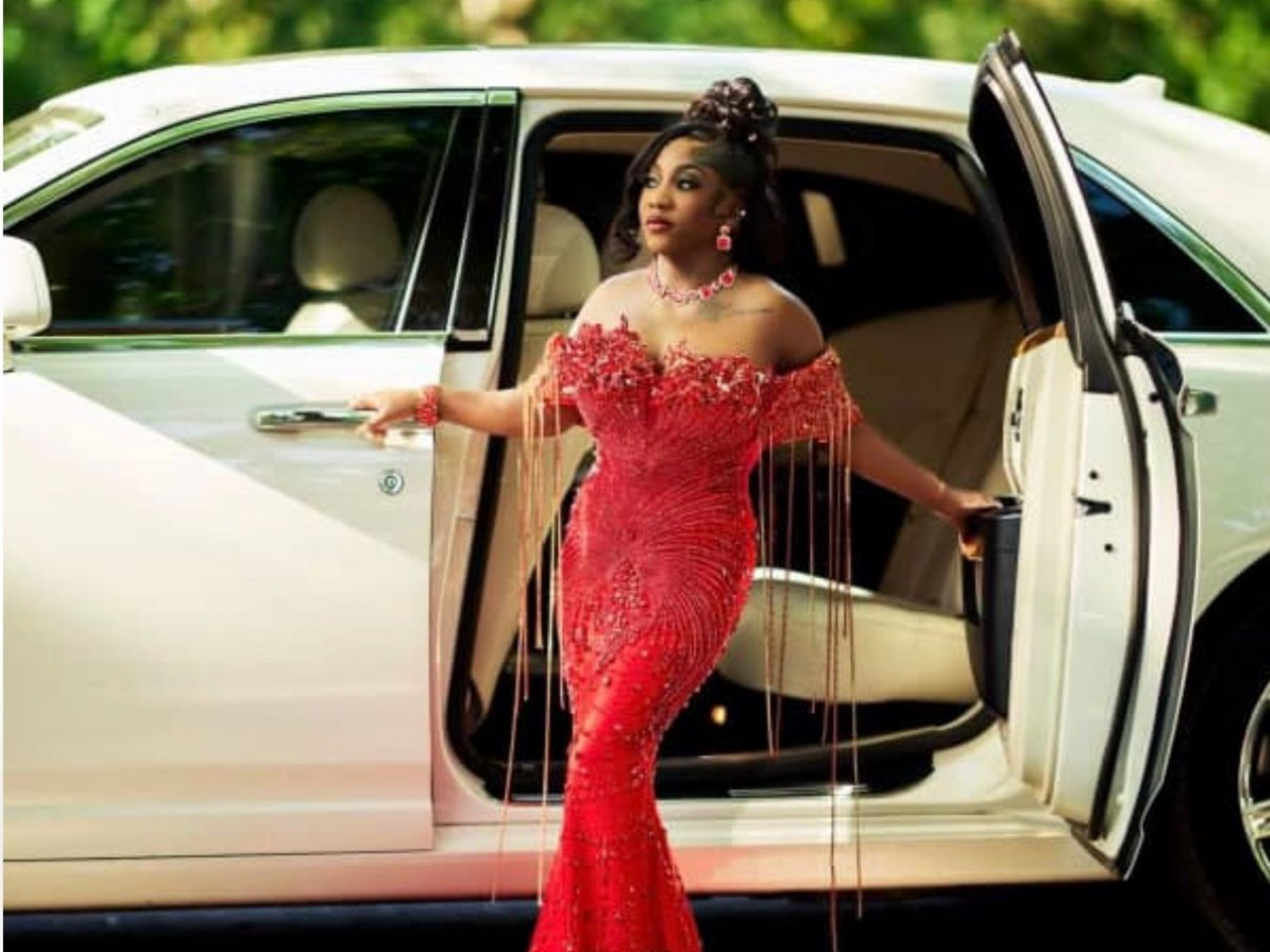 A person in a red, off-the-shoulder beaded gown with fringe accents stands by the open door of a white car.