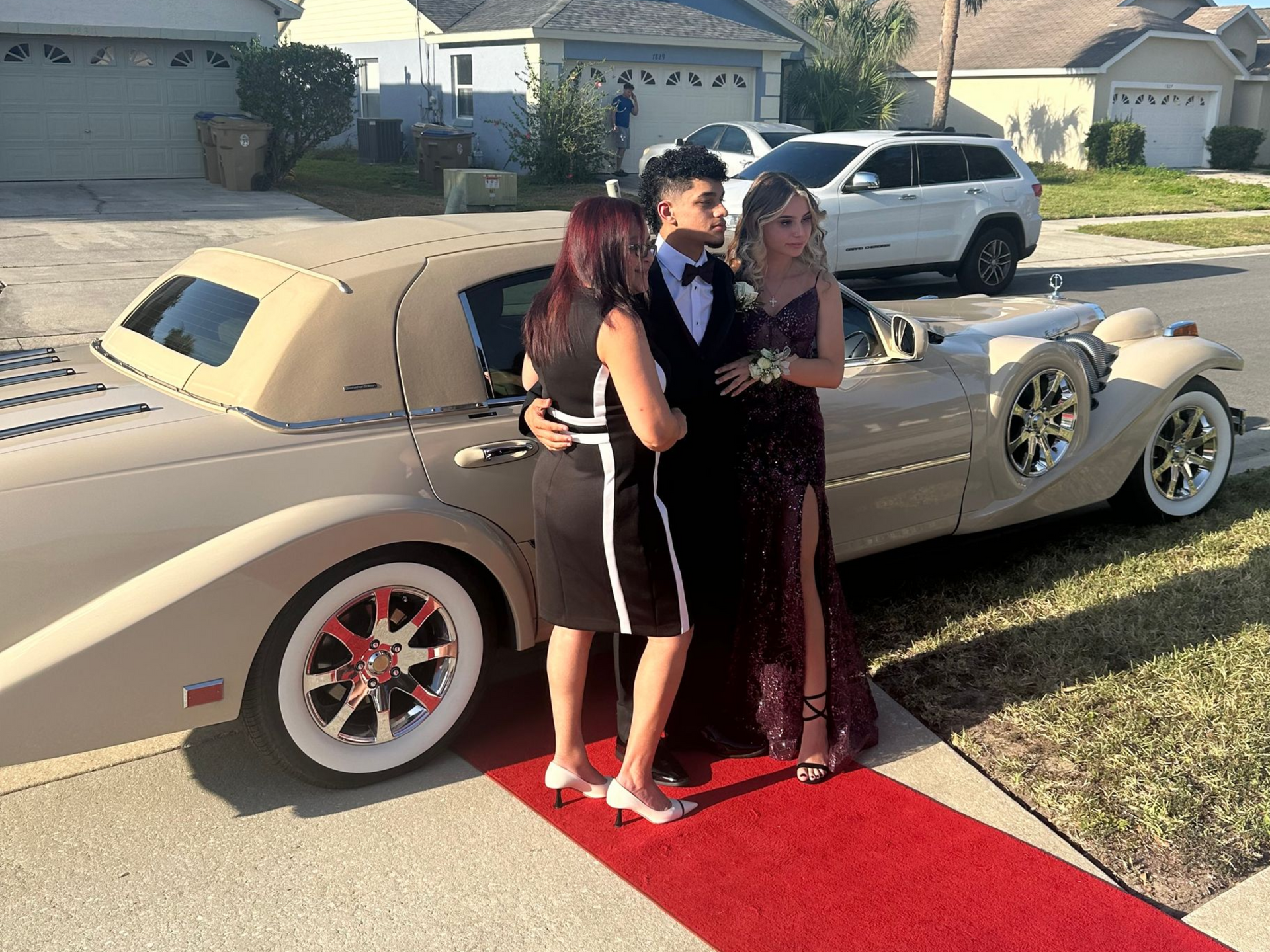 Three people pose on a red carpet next to a vintage-style beige car parked on a suburban street.