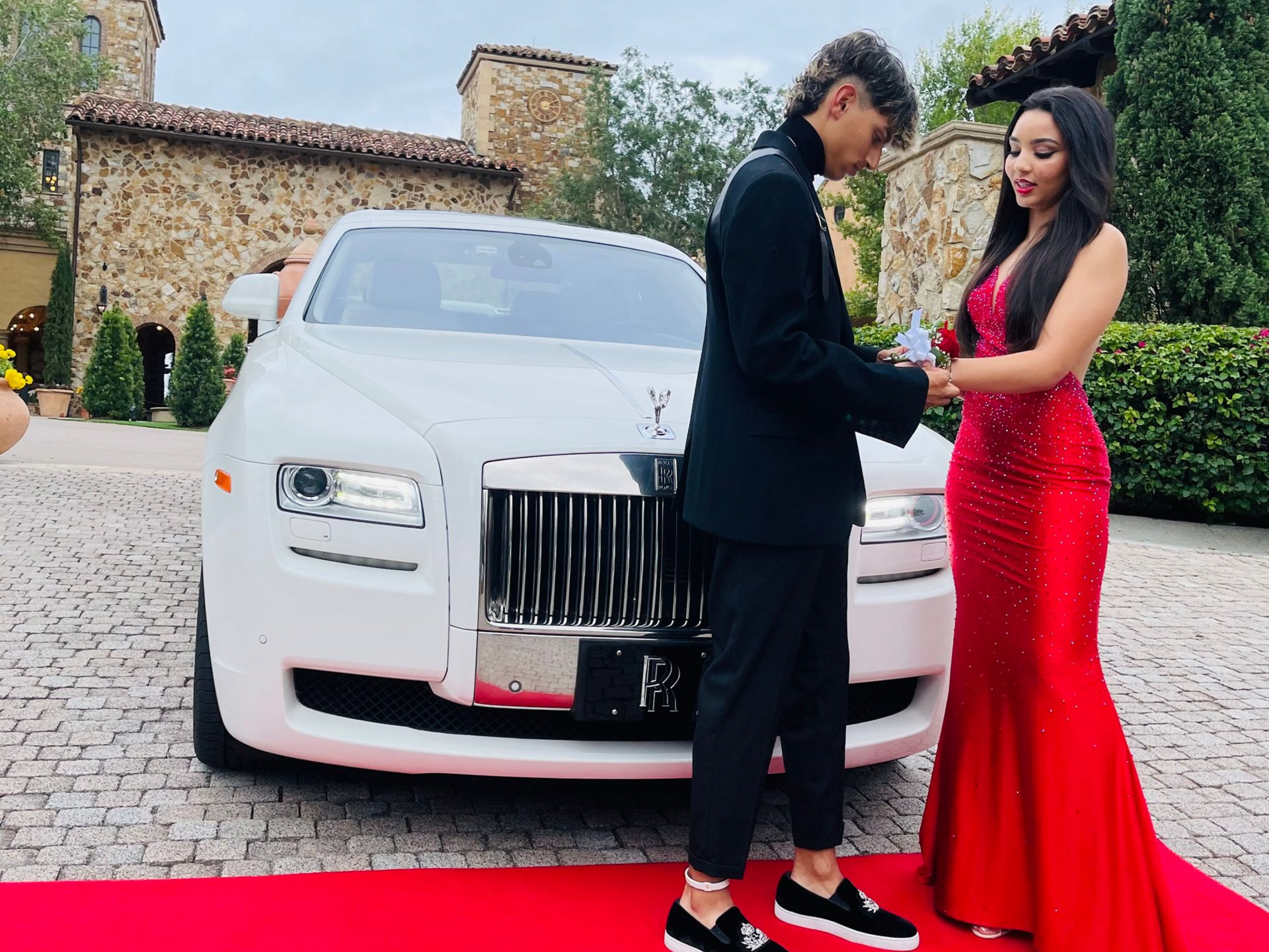 A person in a black suit pins a corsage onto a person in a red gown on a red carpet in front of a white Rolls-Royce.