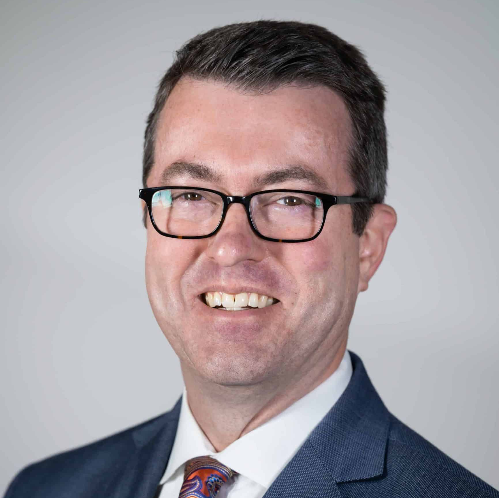 A man wearing glasses and a suit and tie is smiling for the camera.