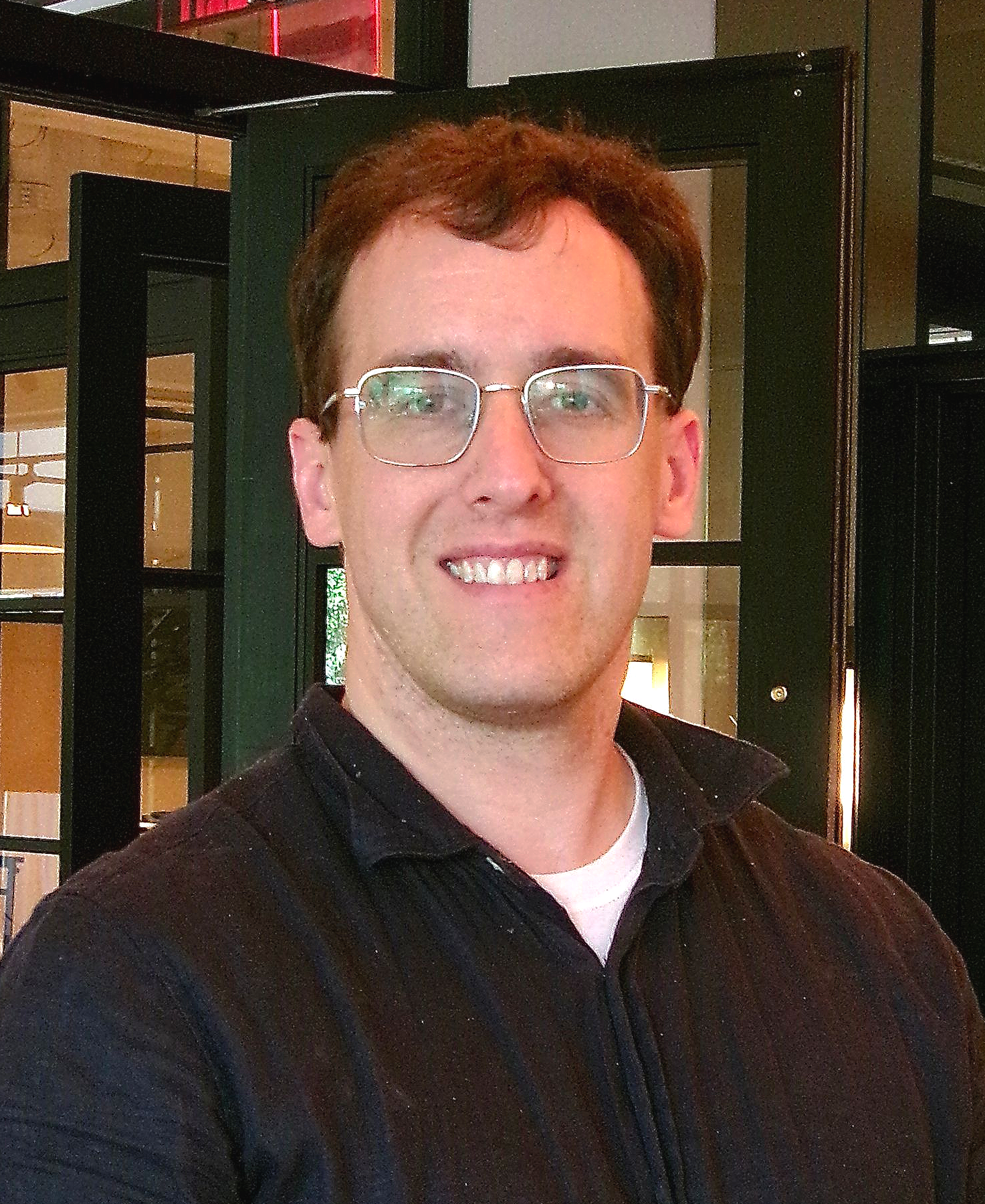 Man in black button up shirt and wire rimmed glasses smiles at camera.