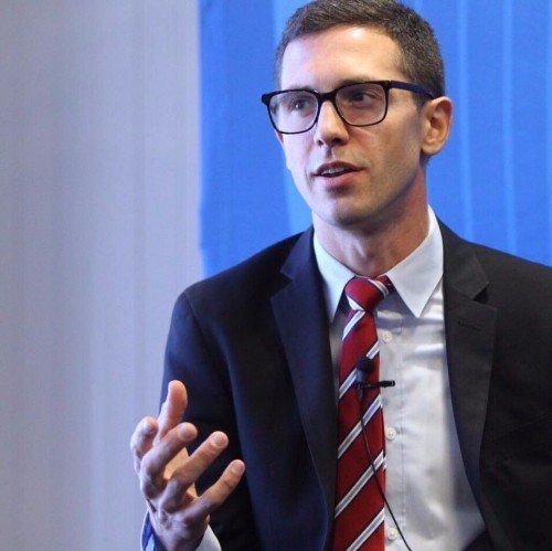 Man in glasses, suit, and red striped tie speaking at a conference, gesturing with hand.