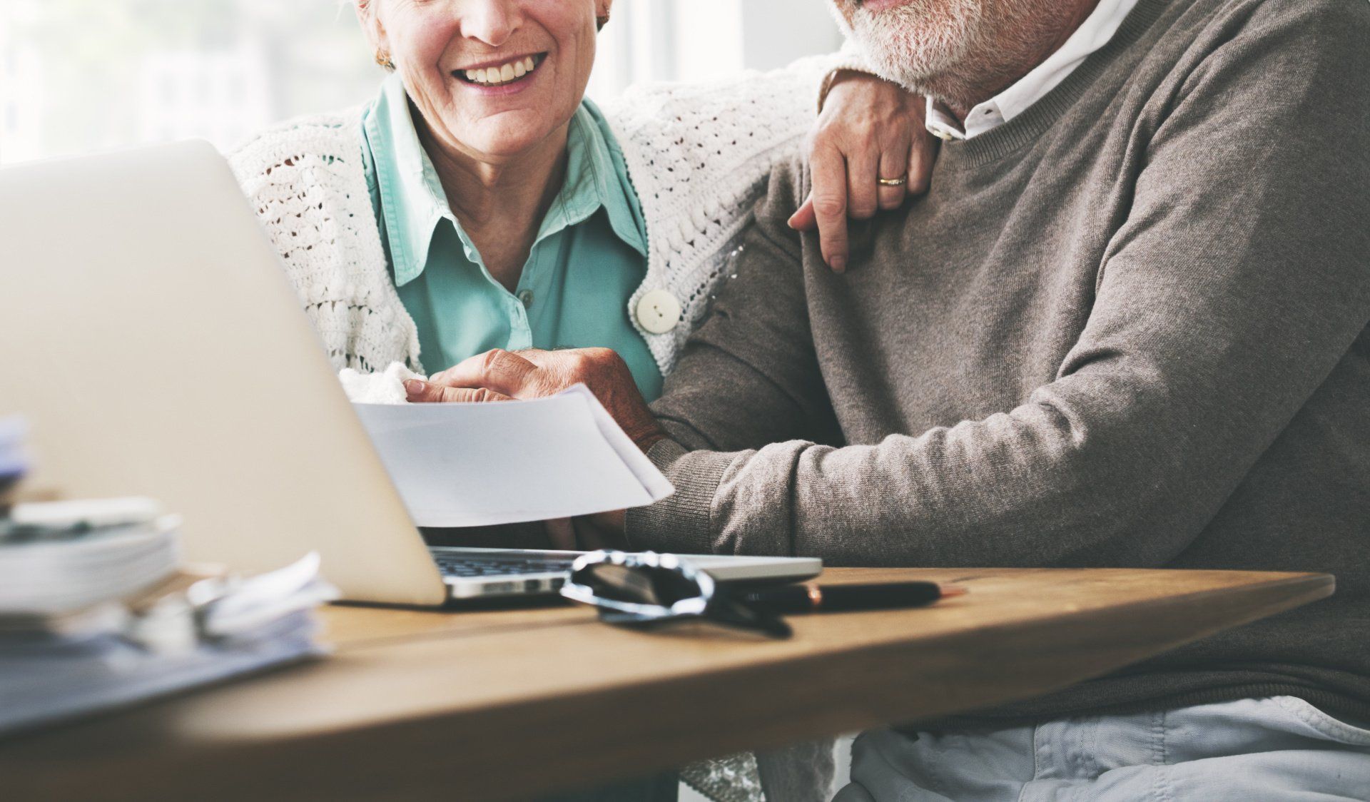 Older couple looks at laptop, smiling, papers on the table.