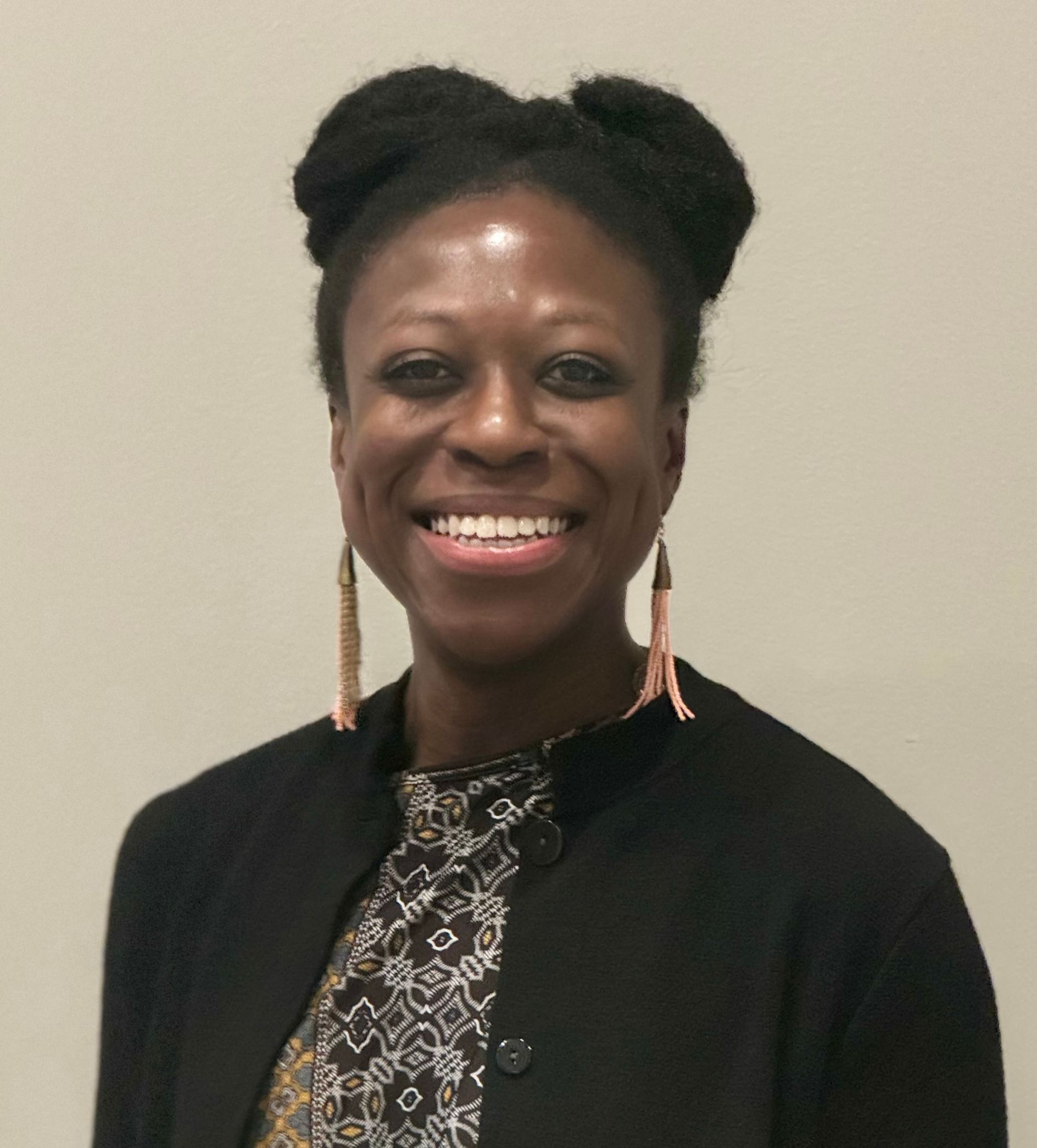 Woman smiling, wearing black jacket and patterned shirt, with two buns and dangling earrings.