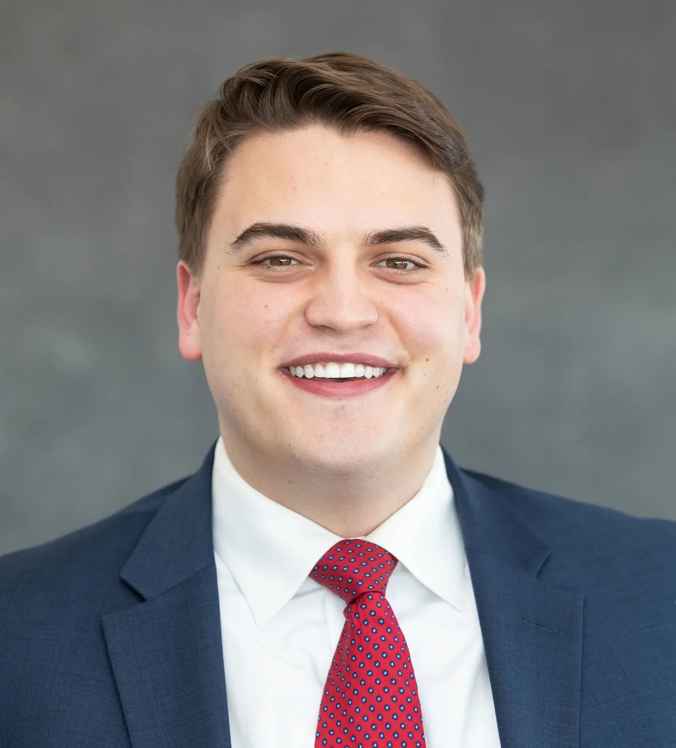 Man in a navy suit and red tie smiles at the camera, against a gray background.