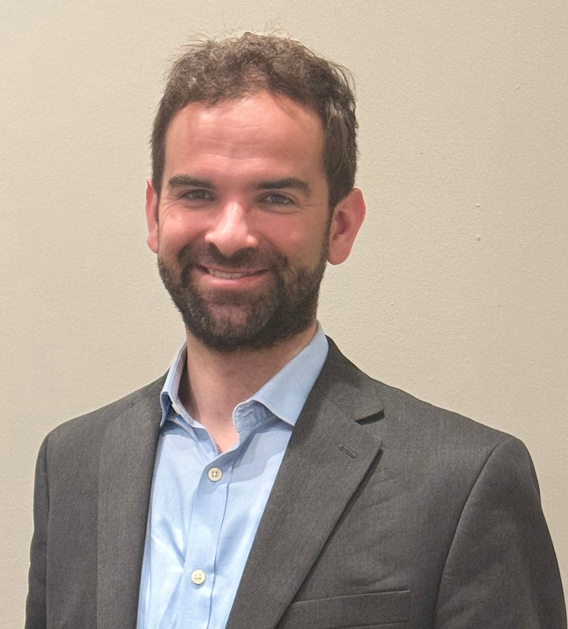 Man with a beard in a blue shirt and dark blazer smiles, against a neutral wall.