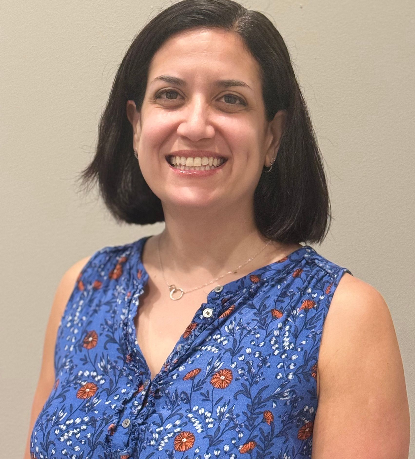 Woman with short dark hair smiling, wearing a blue floral sleeveless top, against a neutral wall.