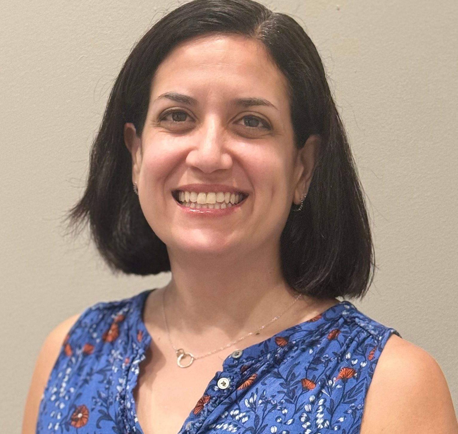 Woman with short dark hair smiling, wearing a blue floral sleeveless top, against a neutral wall.