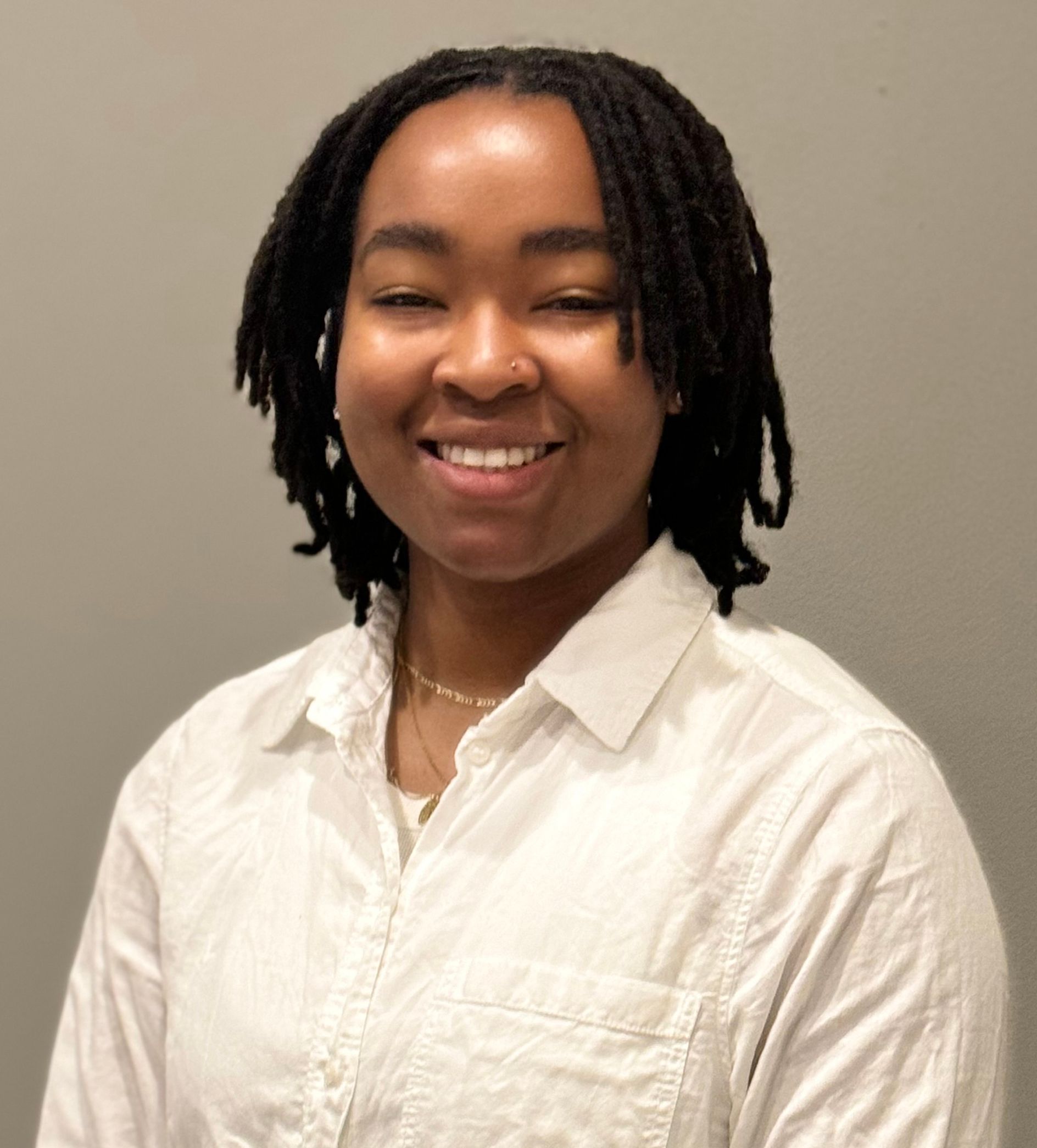 Person with locs smiling, wearing a white button-down shirt, against a gray background.