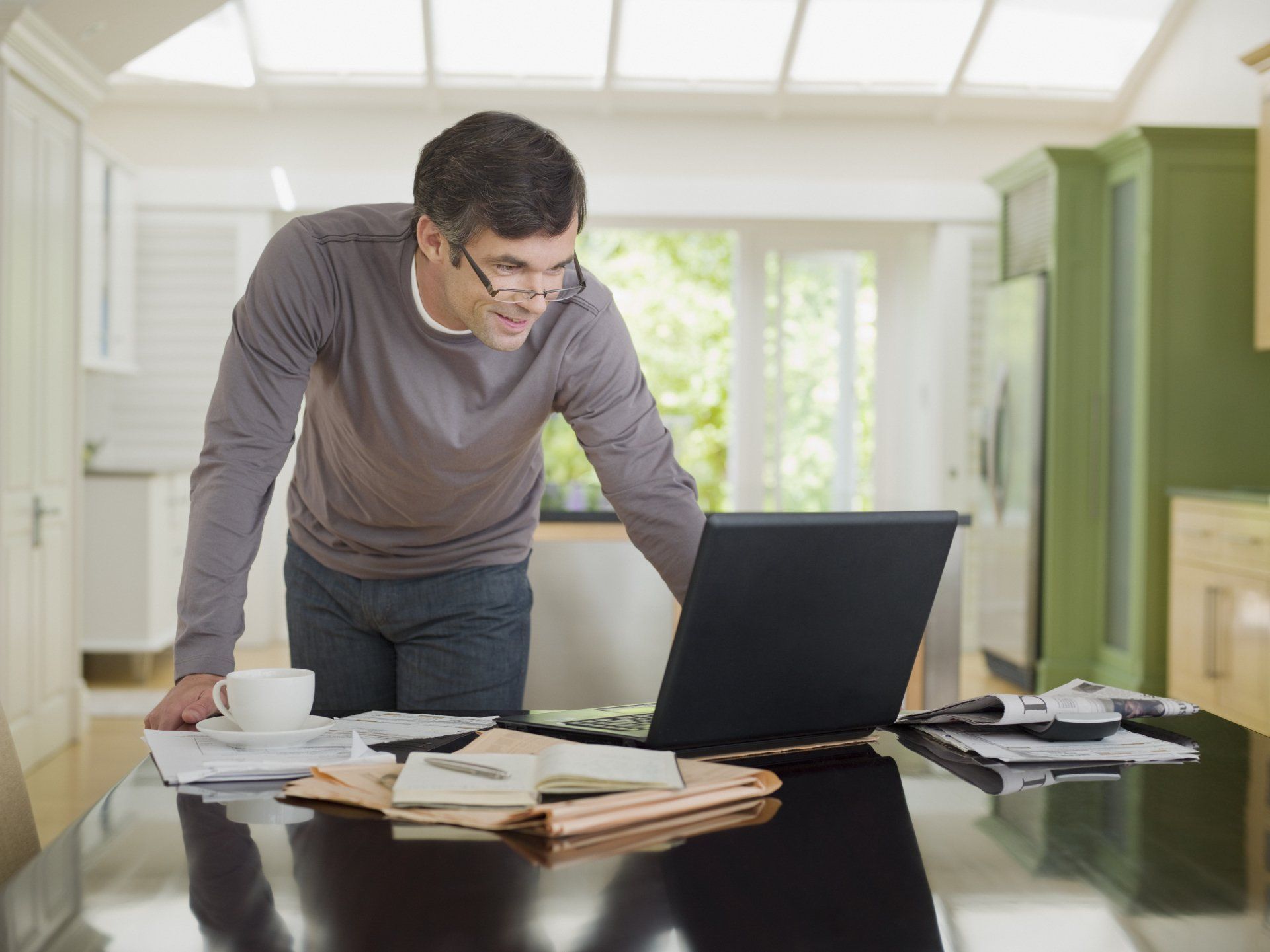Man leans over laptop on table with papers and mug in a kitchen, focused.