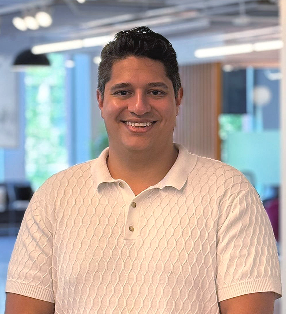 Dark haired man in white textured shirt smiles in front of a blurred office background.