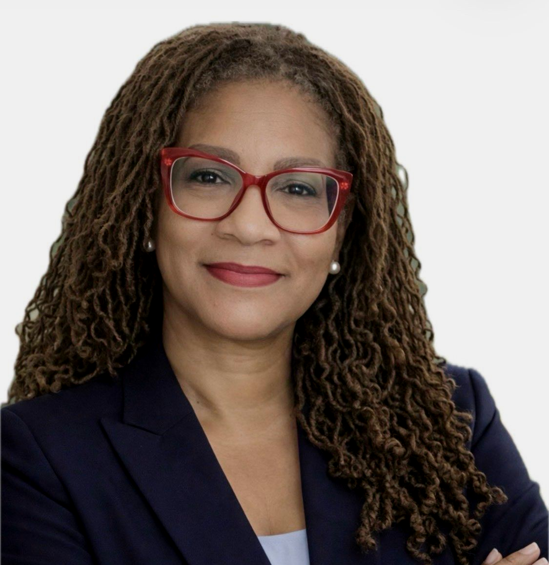 Woman with long, brown locs smiles, wearing red glasses; against a white wall.