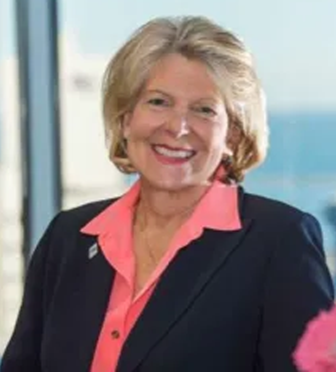 Woman smiling, wearing a pink shirt under a navy blazer. Sunlight from a window illuminates her.