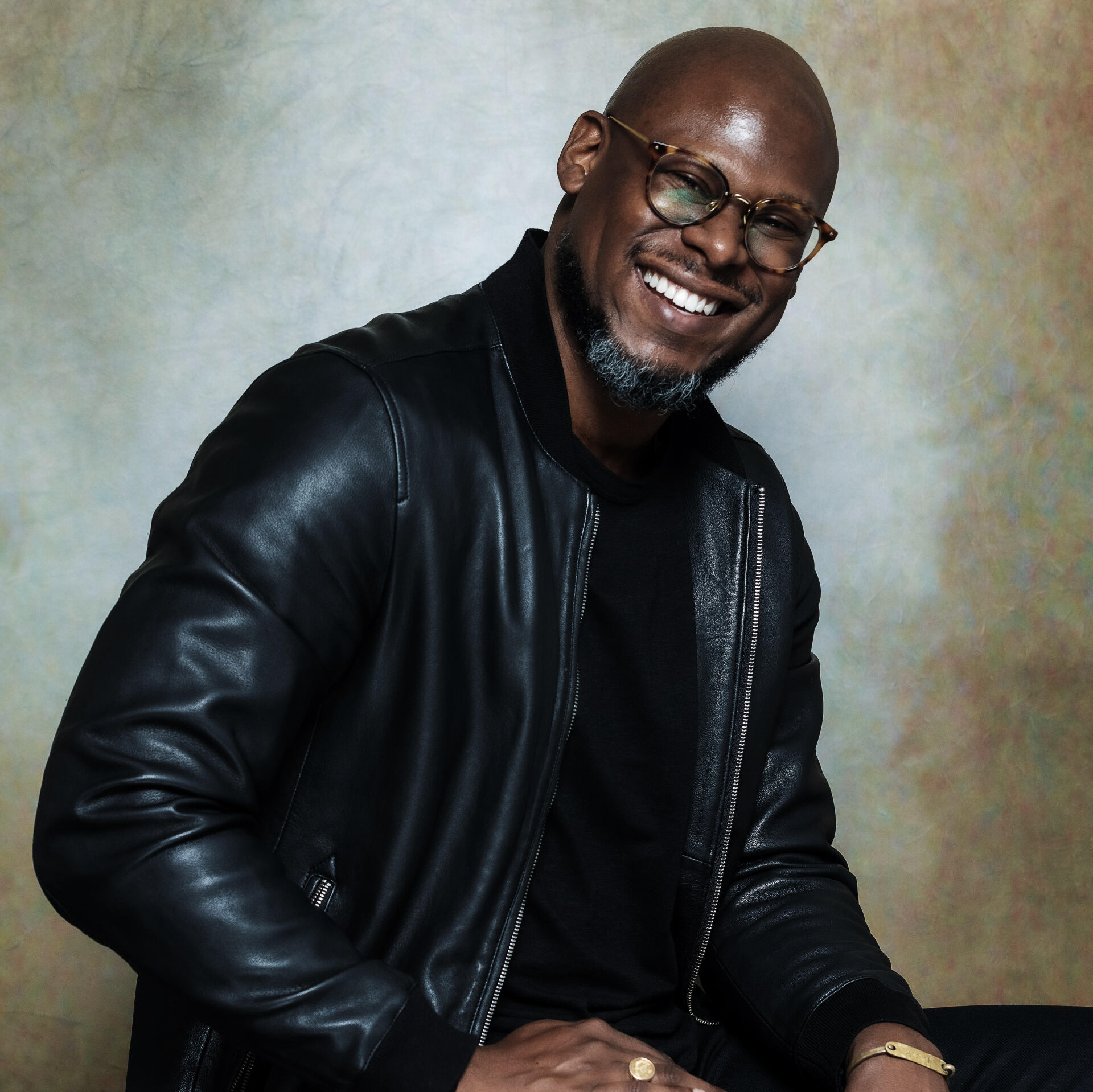 A man wearing glasses and a leather jacket smiles for the camera