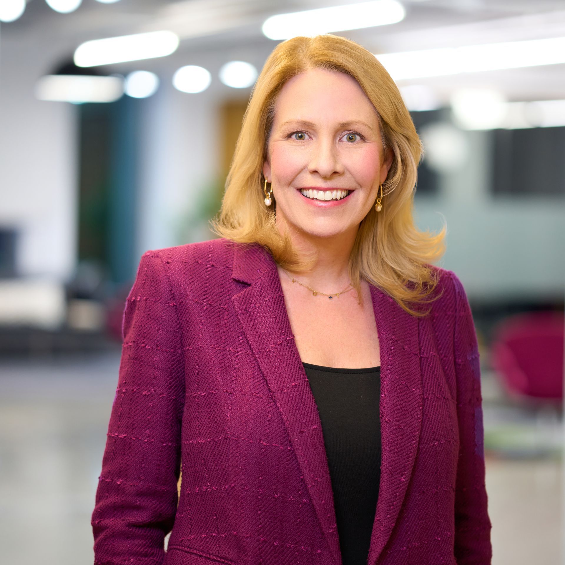 Woman with blonde hair smiles, wearing a light blue sweater and necklace, gold earrings, background of gold balloons.