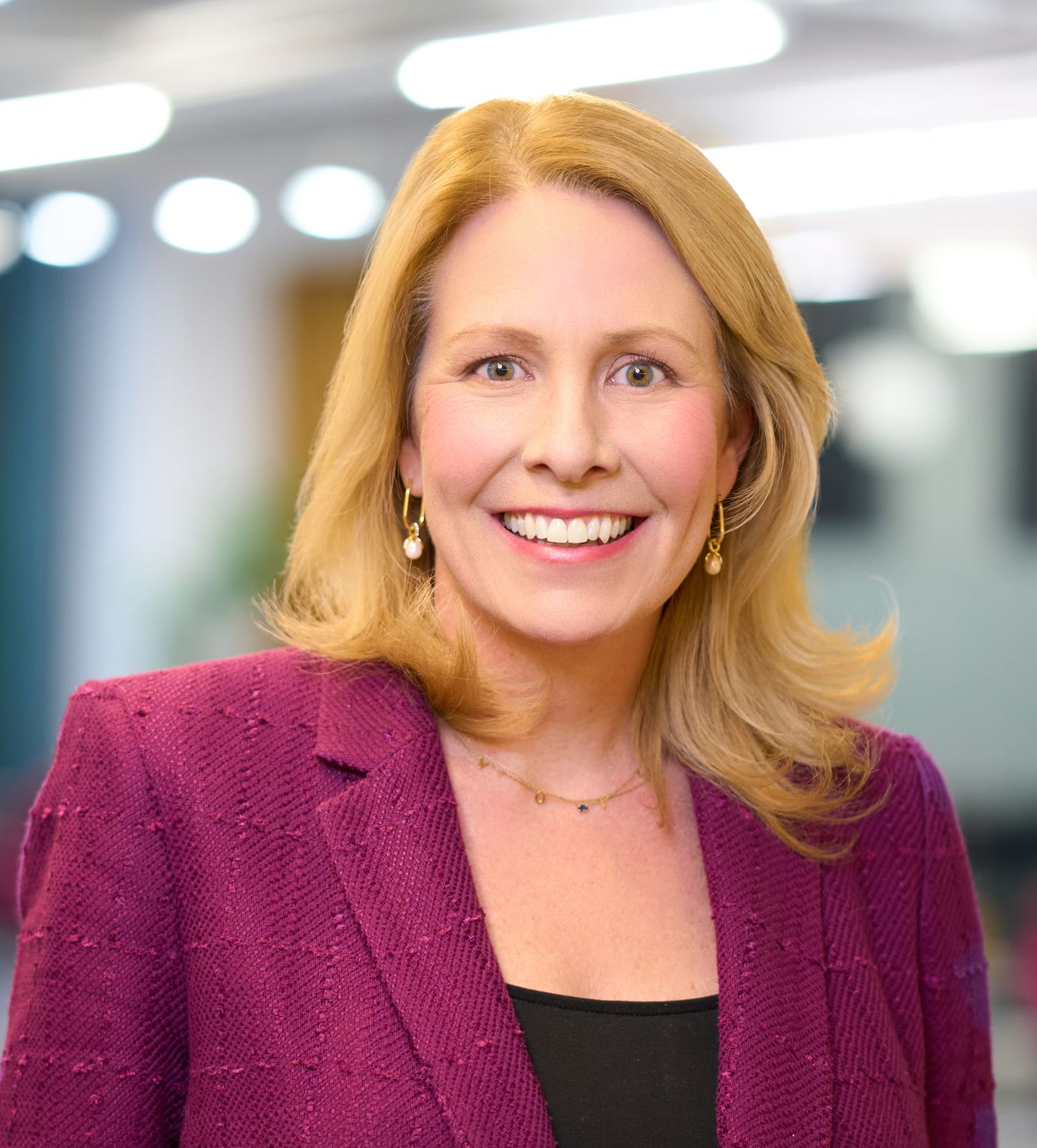 Woman with blonde hair, smiling, wearing a purple blazer and gold earrings, against a blurred office background.