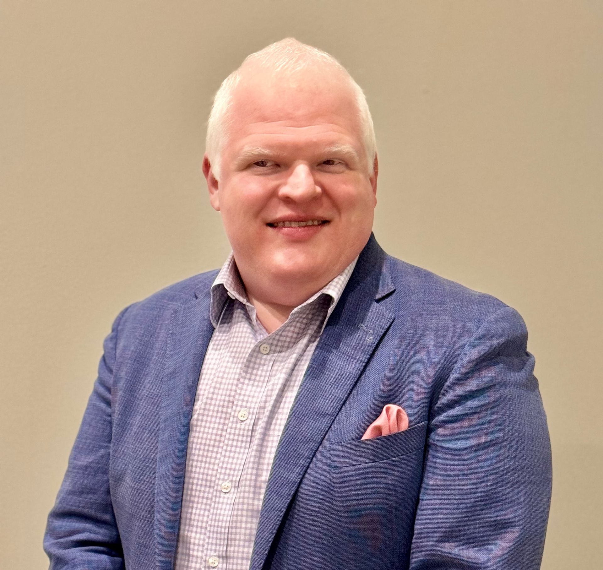 Smiling man in blue blazer and patterned shirt, pocket square visible, indoors.