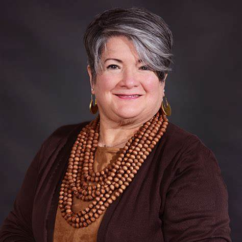 A woman with gray hair is wearing a brown sweater and a wooden necklace.