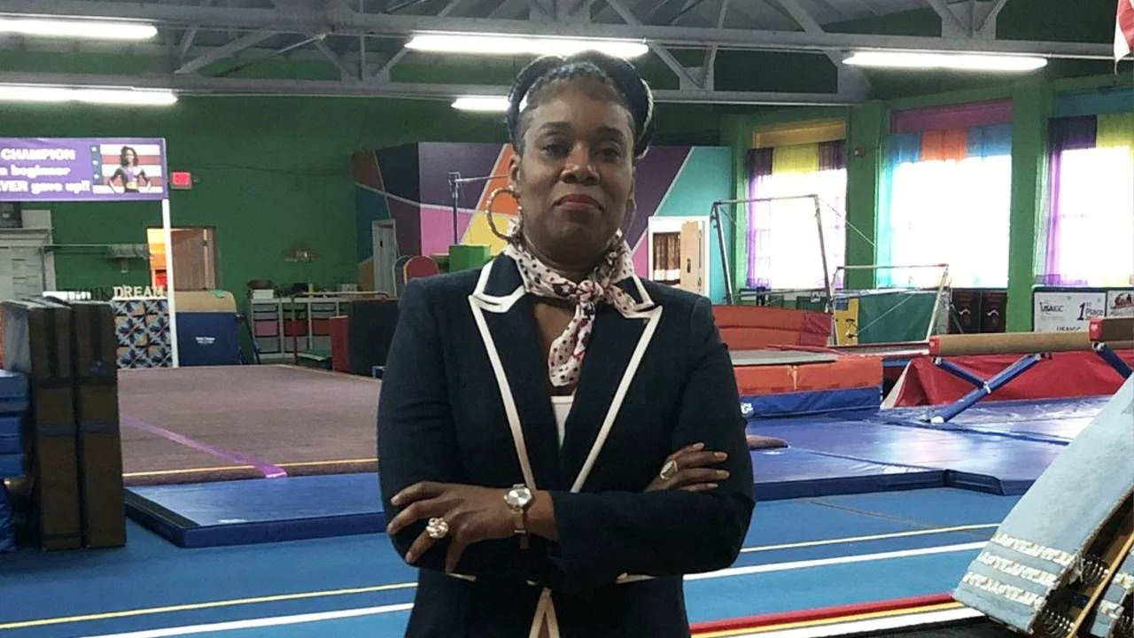 Woman in blazer with arms crossed, in gymnastics studio with mats and equipment.