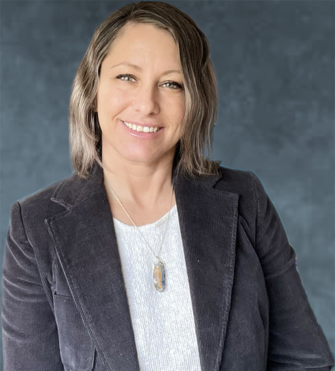 Woman smiling, wearing a white shirt, gray corduroy blazer, and pendant necklace. Dark blue backdrop.