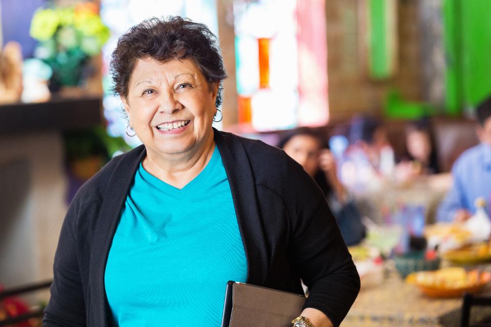Woman in teal shirt and black sweater smiles, holding tablet in a restaurant.