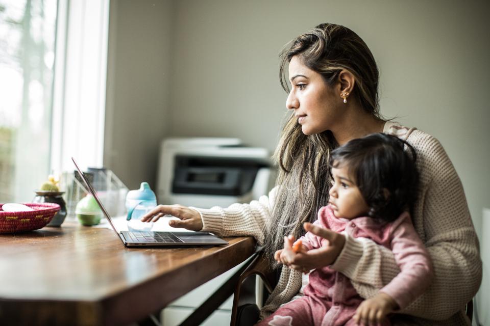 Woman using laptop with a toddler on her lap, sitting at a table near a window.